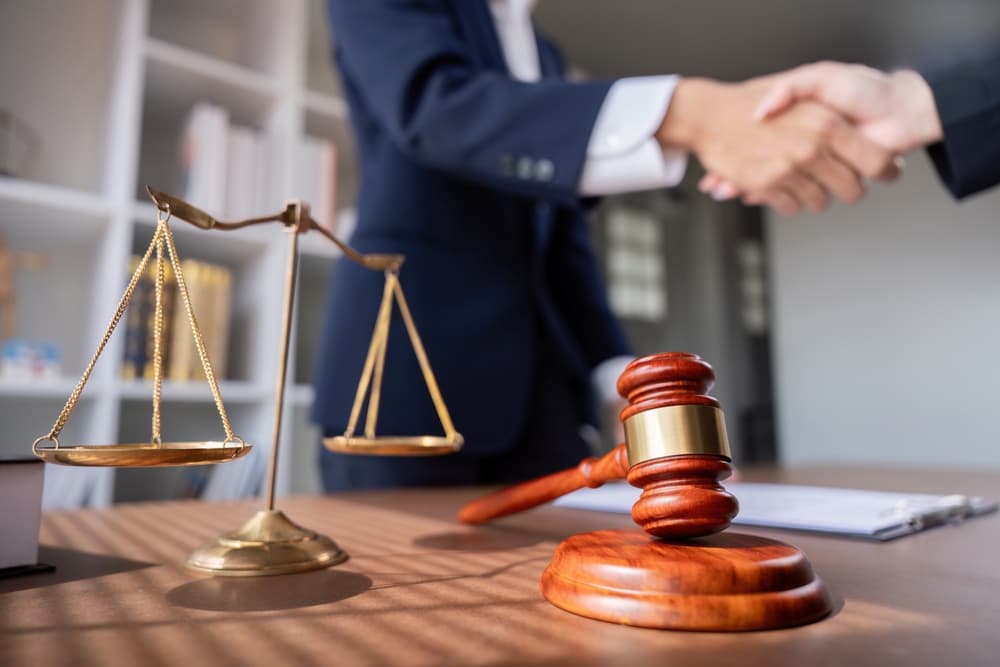 A professional lawyer's handshake during a business meeting, with scales of justice and a gavel on the desk, symbolizing a legal agreement and partnership in a modern office setting.